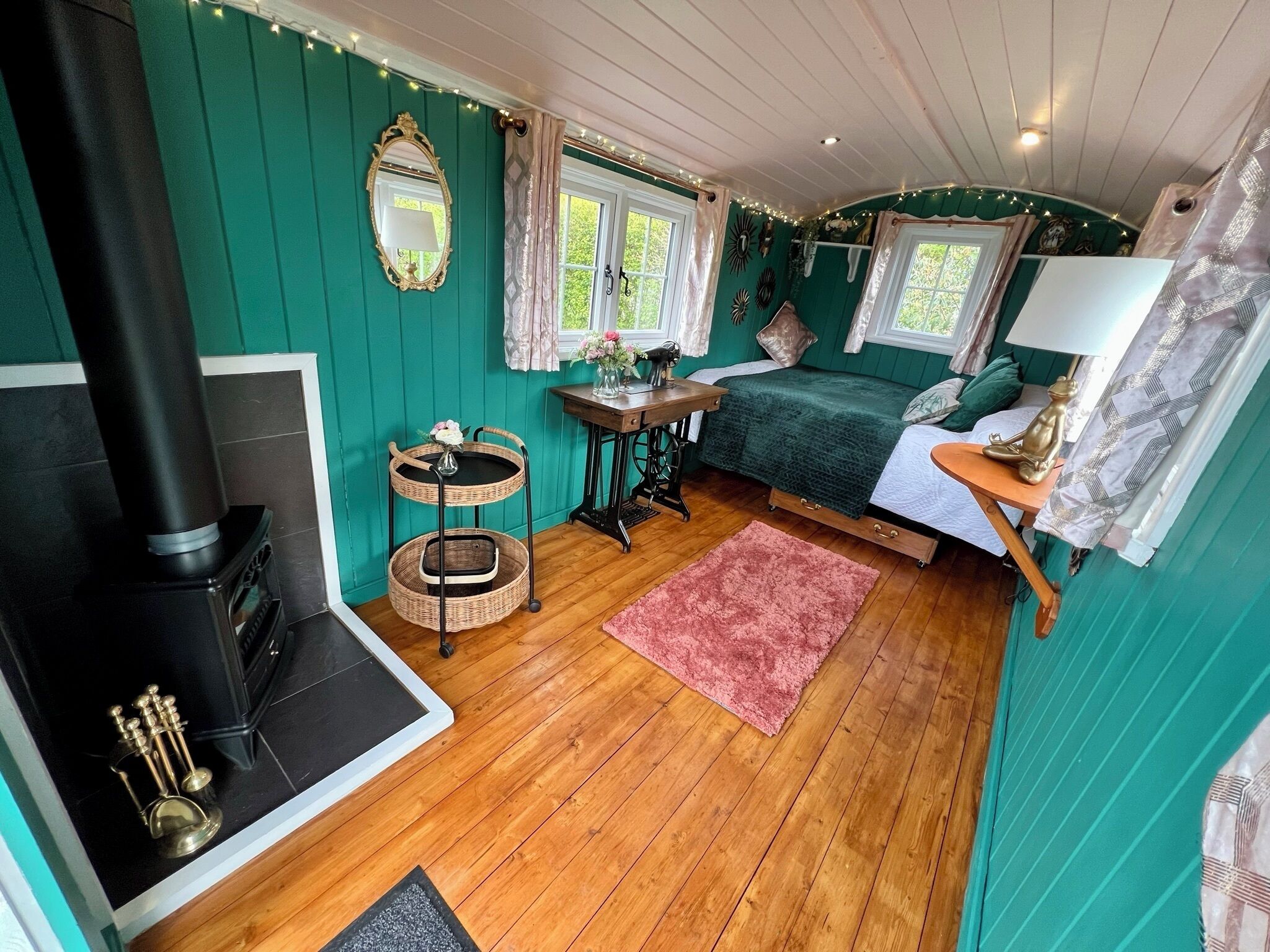 A cozy room with a bed, a small table, a sewing machine, a wicker shelf, and a fireplace.