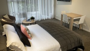 Ett modernt hotellrum med en säng, ett litet runt bord med en växt, ett skrivbord med två stolar och en tv på väggen.