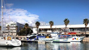 Un puerto deportivo con varias embarcaciones amarradas, un edificio con la señalización 'MADE DE LAGO' y palmeras al fondo.