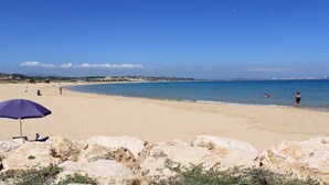 Una playa con un primer plano rocoso, cielo azul despejado y algunas personas a lo lejos.