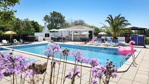 Un área de piscina con sillones de descanso, un pabellón y un edificio blanco con techo a dos aguas.