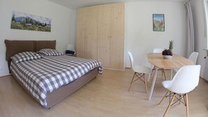 A bedroom with a bed, a wooden wardrobe, a dining table with chairs, and two paintings on the walls.