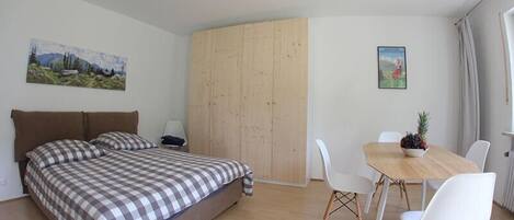 A bedroom with a bed, a wooden wardrobe, a dining table with chairs, and two paintings on the walls.