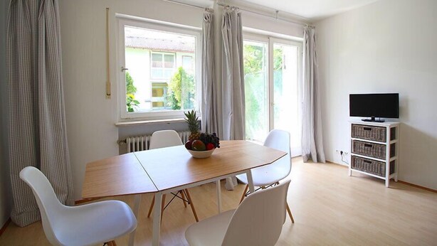 A bright room with a wooden dining table, white chairs, a fruit bowl, a TV on a white stand, and large windows with curtains.