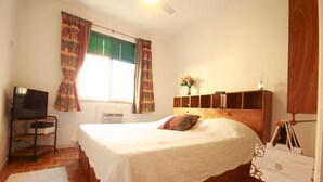 Ein Schlafzimmer mit Bett, Fernseher auf einem Schrank, Fenster mit Vorhängen, einem Holz-Kopfteil, einem Nachttisch mit Vase und einem weißen Teppich.
