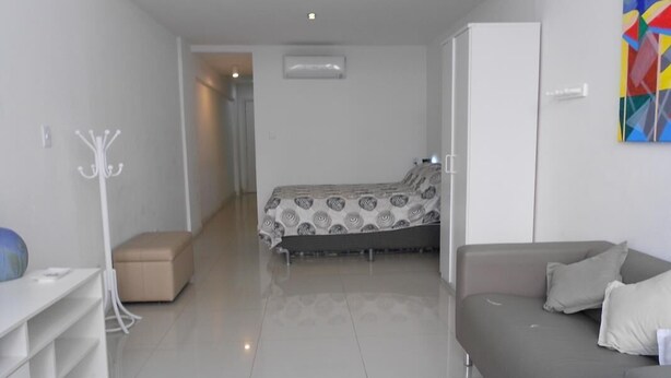 A modern, well-lit room with a bed, a grey sofa, a white coat rack, and a colorful abstract painting on the wall. - Gohouse - Copacabana 1206 (Rio de Janeiro)