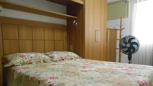 A bedroom with a floral bedspread, wooden headboard, wardrobe, and a standing fan. - Gohouse - Dutra 102 (Rio de Janeiro)
