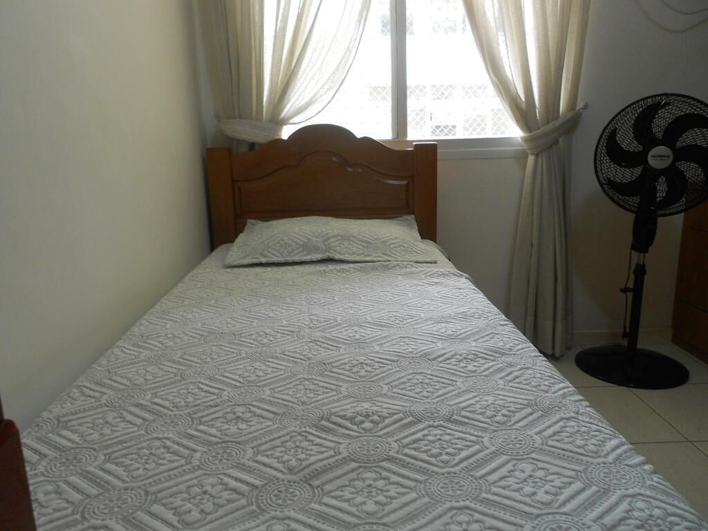 Una cama individual con un edredón estampado, un cabecero de madera y un ventilador de pie a la derecha.