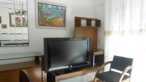 A modern living room with a flat-screen TV on a wooden stand, a painting on the wall, and a window with curtains. - Gohouse - Dutra 102 (Rio de Janeiro)