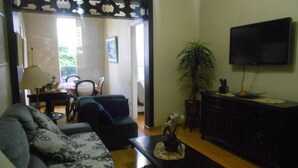 A living room with a sofa, a coffee table, a TV, a plant, and a dining area in the background. - Gohouse - Figueiredo 601 (Rio de Janeiro)