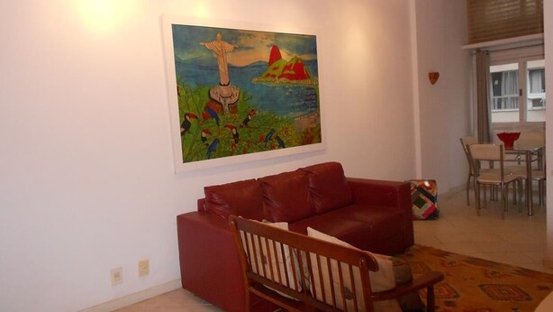 A living room with a red sofa, a wooden chair, a painting on the wall, and a dining area with chairs and a table. - Gohouse - Figueiredo 303 (Rio de Janeiro)