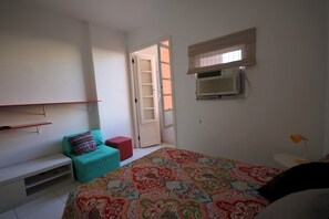 A bedroom with a bed, a small seating area, a window with blinds, and an air conditioning unit. - Gohouse - Raul 607 (Rio de Janeiro)
