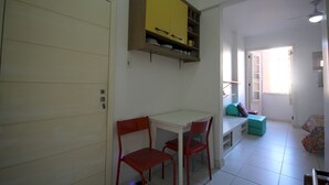 Ein kleiner, heller Raum mit einer weißen Tür, einem gelben Schrank über einem weißen Tisch mit zwei roten Stühlen und einem Fenster mit Blick nach draußen.