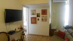 A living room with a television, a sofa, a small table, and a chair. There are wall decorations and a window with curtains. - Gohouse - Raul 1003 (Rio de Janeiro)