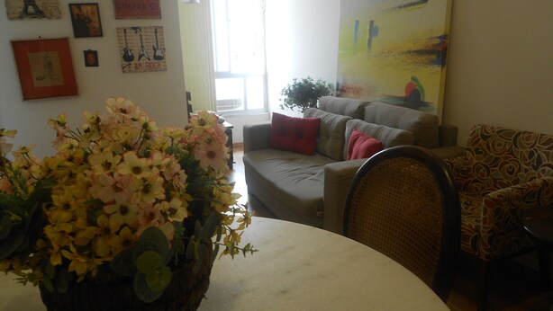 A living room with a sofa, a patterned armchair, a table with a floral arrangement, and various wall decorations including posters and a painting. - Gohouse - Raul 1003 (Rio de Janeiro)