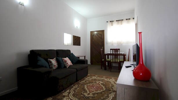 A living room with a sofa, a coffee table, and a red vase. There is a door and a window with curtains. - Gohouse - Rangel SP 96 (São Paulo)