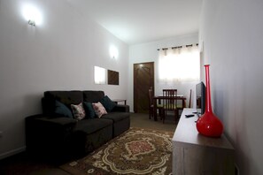 Un salon avec un canapé, une table basse et un vase rouge. Il y a une porte et une fenêtre avec des rideaux.