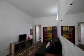 Un salon avec un téléviseur, un vase rouge, un tapis à motifs et un canapé. On aperçoit des étagères colorées à l’arrière-plan.