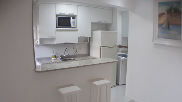 A modern kitchen with white cabinets, a microwave, a sink, and a dishwasher. There are two white barstools and a painting on the wall. - Gohouse - Torre do Barao 108 (Rio de Janeiro)