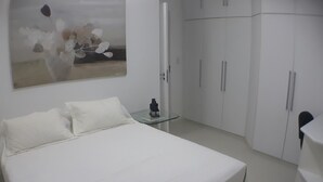 A bedroom with a large bed, white walls, and a painting of flowers. There are white cabinets and a small table with a remote control. - Gohouse - Torre do Barao 108 (Rio de Janeiro)