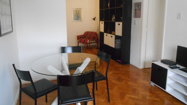 A dining area with a glass table and black chairs, a black cabinet with shelves, and a red chair with a striped blanket. - Gohouse - Torre 302 C (Rio de Janeiro)