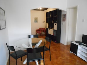 A dining area with a glass table and black chairs, a black cabinet with shelves, and a red chair with a striped blanket.