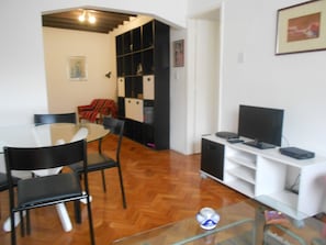 A dining area with a glass table and chairs, a television on a white stand, and a bookshelf in the background.
