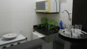 Una cocina con microondas, pileta y cocina. Hay sillas verdes, una bandeja con vasos y una botella de detergente.