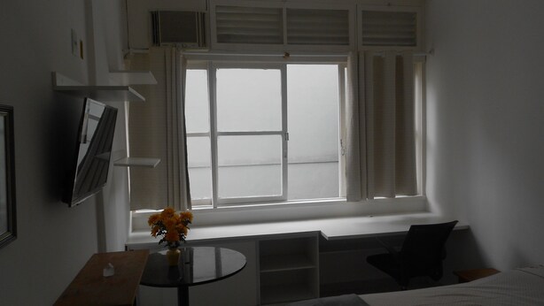 Habitación de hotel con una cama, una mesita con un jarrón de flores, una ventana con cortinas y un televisor en la pared.