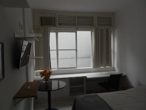 A hotel room with a bed, a small table with a vase of flowers, a window with curtains, and a television mounted on the wall.