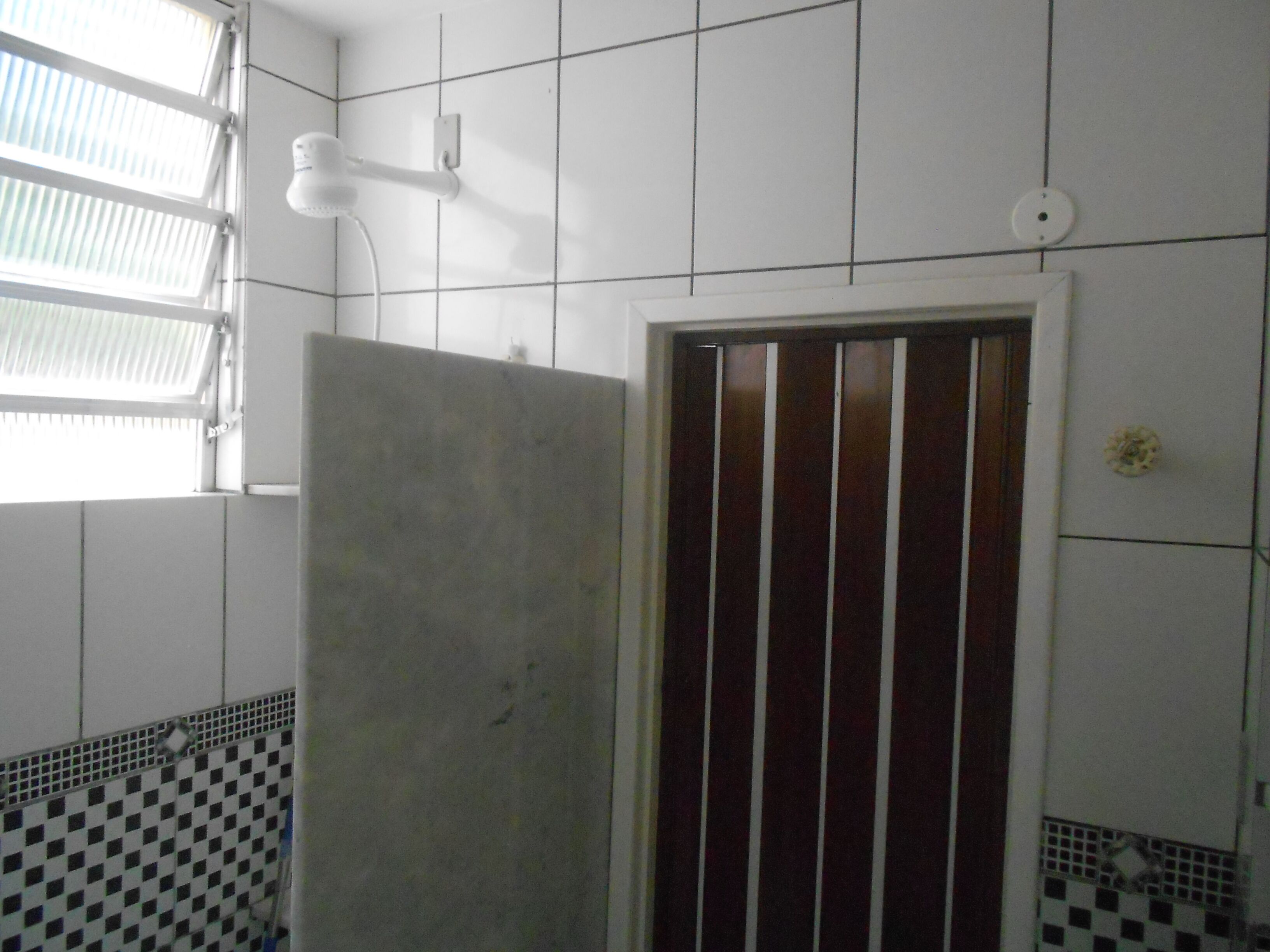 A bathroom with white tiled walls, a checkered tile border, a shower area, and a window with blinds.