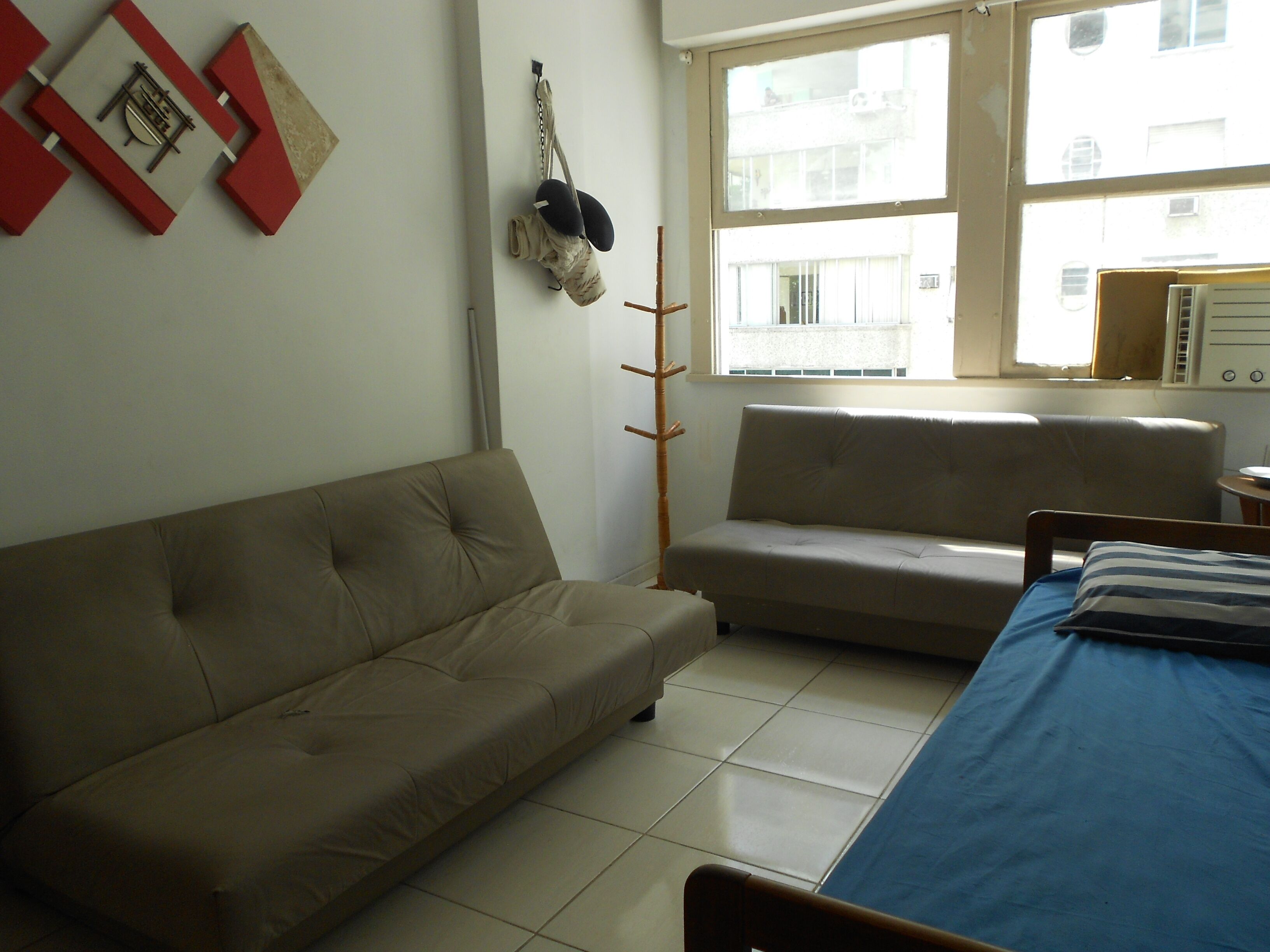 Une pièce avec un canapé beige, une couvre-lit bleue et une fenêtre donnant sur des immeubles.