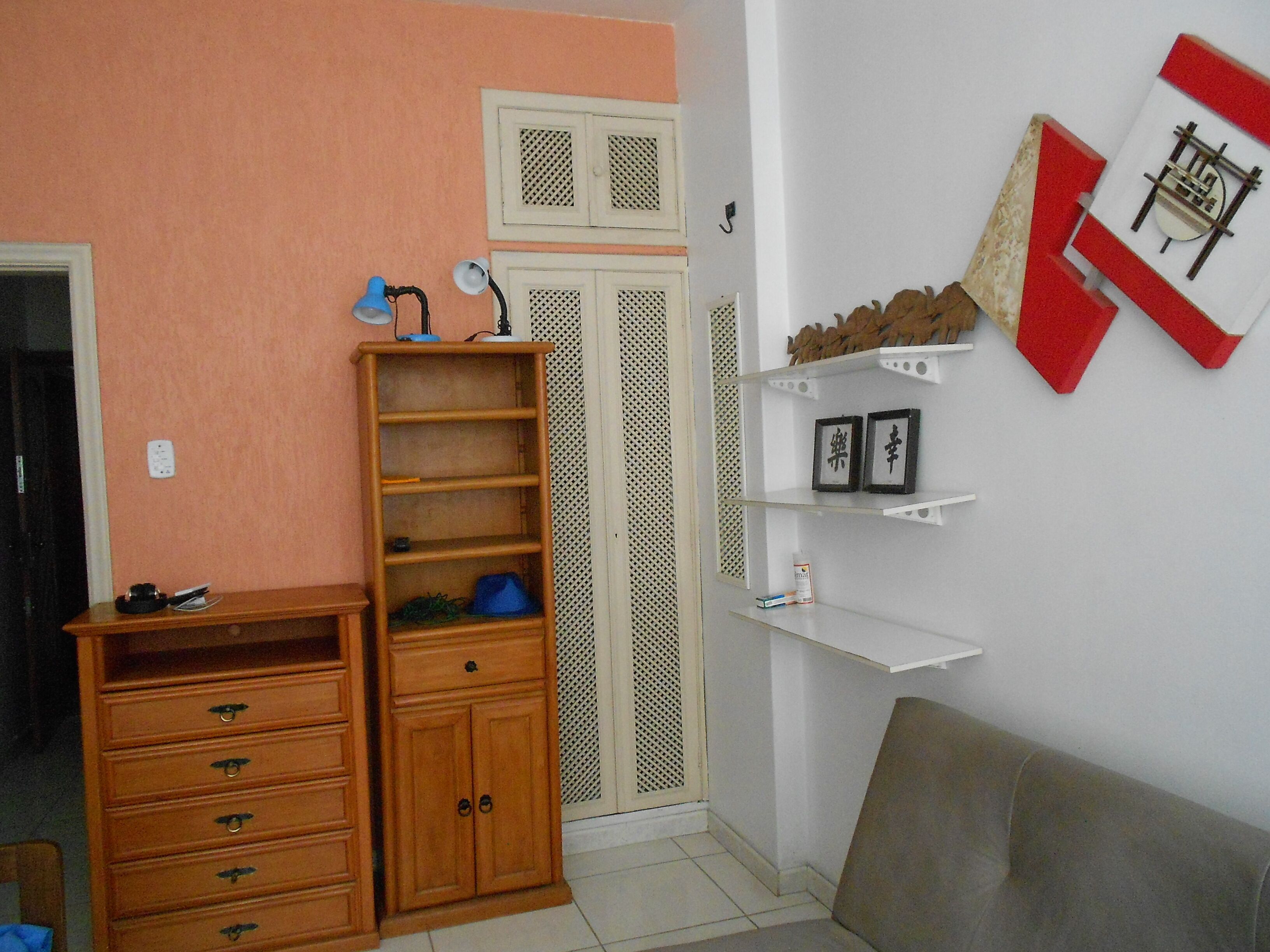 Una habitación con un aparador de madera, una lámpara azul, un estante blanco con fotografías enmarcadas y un adorno rojo y blanco en la pared.