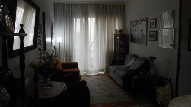 A dimly lit living room with a sofa, a small table with a vase, and a large window with curtains. - Gohouse - Loureiro 102 (Rio de Janeiro)