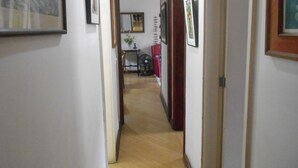 A narrow hallway with wooden flooring and white walls, leading to a room with a bed and a chair. - Gohouse - Loureiro 102 (Rio de Janeiro)