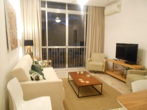 Una sala de estar con un sofá, sillones, una mesa de centro, un televisor sobre un mueble de madera y un amplio ventanal con cortinas.