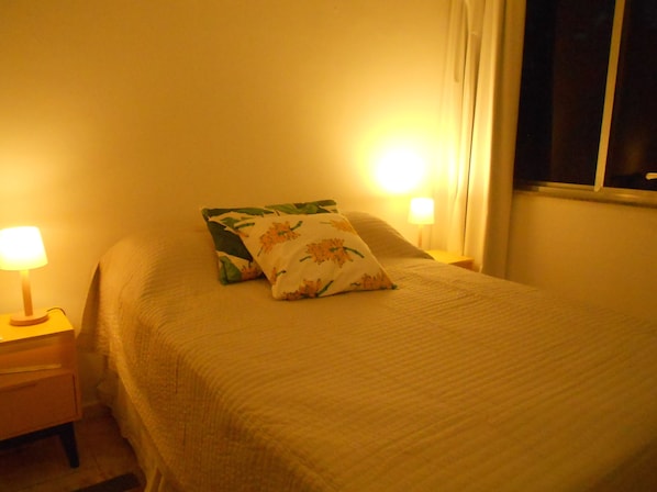 A neatly made bed with floral pillows, a bedside lamp, and a window with curtains. - Gohouse - Elizabeth 305 (Rio de Janeiro)