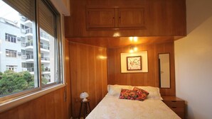 Un dormitorio con cama, mesita de noche, lámpara, un cuadro enmarcado en la pared y ventana con vistas a edificios y árboles.