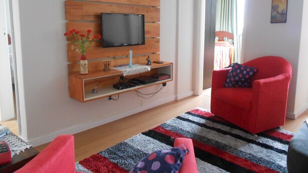A living room with a flat-screen TV mounted on a wooden panel, a red armchair, a striped rug, and a small table with a vase of flowers. - Gohouse - Sacadura 601 (Rio de Janeiro)