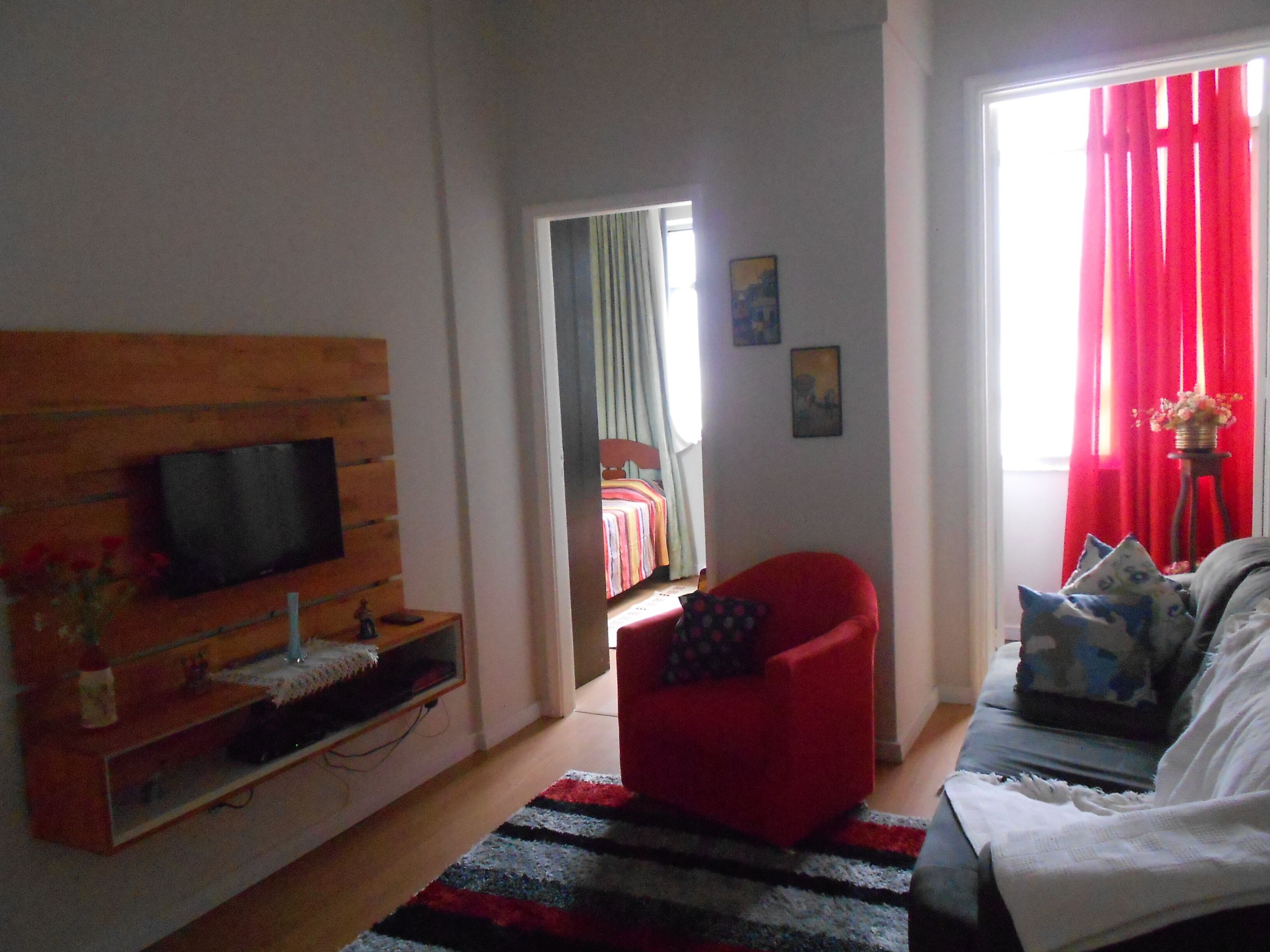 A living room with a TV mounted on a wooden panel, a red armchair, a sofa with patterned cushions, and a view of a bedroom through an open door.