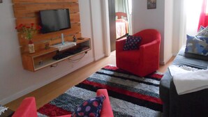 A modern living room with a flat-screen TV mounted on a wooden panel, a red armchair, a patterned rug, and a bed with a colorful bedspread in the background. - Gohouse - Sacadura 601 (Rio de Janeiro)