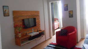 A living room with a flat-screen TV mounted on a wooden panel, a red armchair, and a small table with a vase of flowers. - Gohouse - Sacadura 601 (Rio de Janeiro)