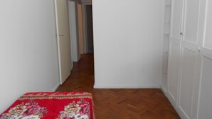 A narrow hallway with wooden flooring, white walls, and a red floral bedspread. - Gohouse - Siqueira 1003 (Rio de Janeiro)
