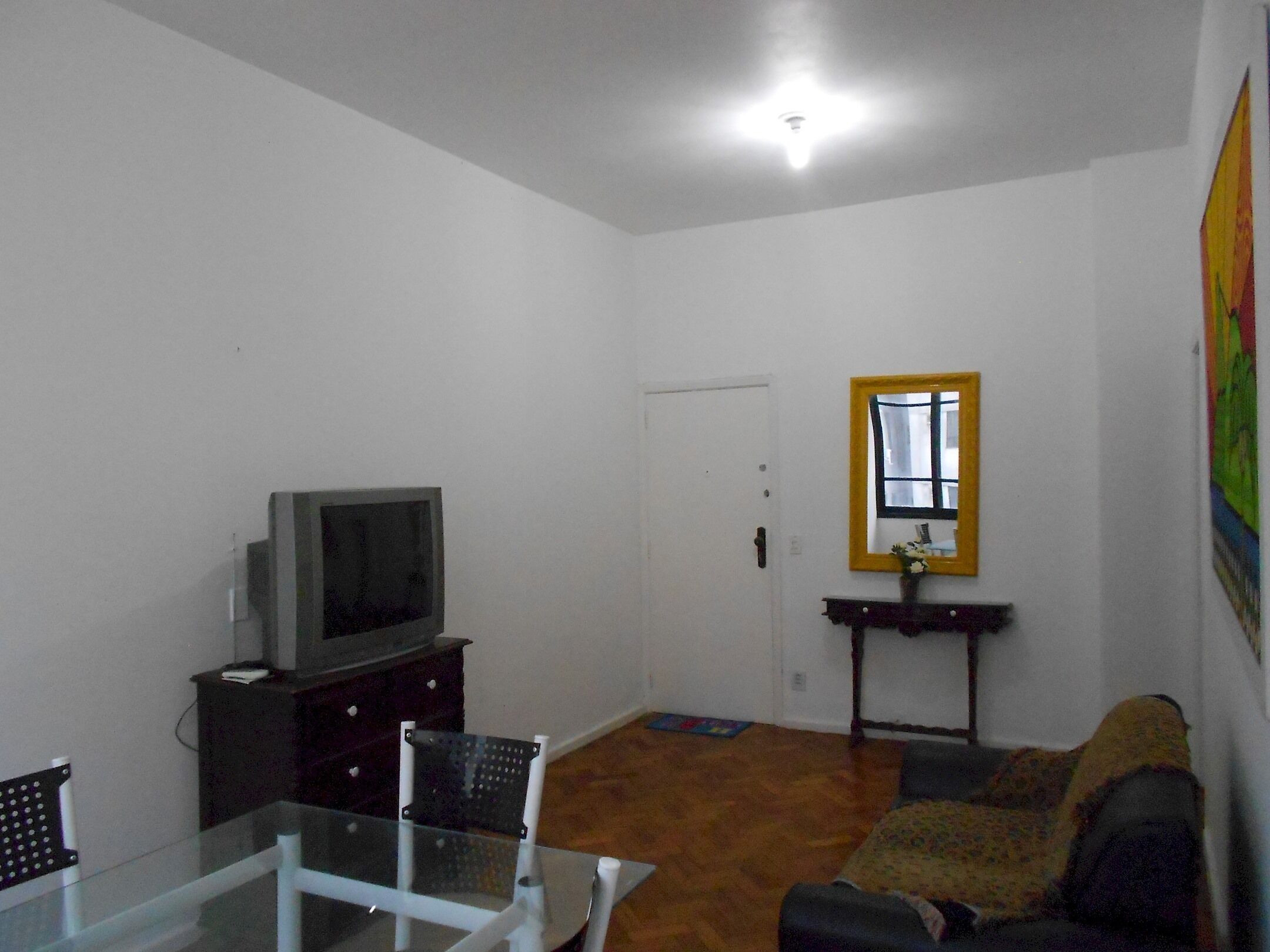 Uma sala de estar com televisão sobre um suporte, uma mesa de vidro, um sofá, uma poltrona e uma porta.