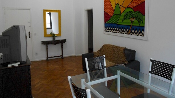 A living room with a glass dining table, a sofa, a television, and a colorful painting on the wall. - Gohouse - Siqueira 1003 (Rio de Janeiro)