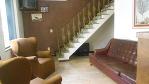 Un salon avec un canapé en cuir marron, un fauteuil marron et un canapé rouge. On aperçoit un escalier avec une rampe dorée au fond.
