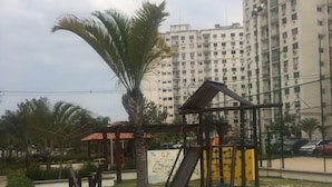 A playground with a slide and swings, surrounded by greenery and a tall building in the background.