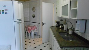 A compact kitchen with a refrigerator, a dining table with chairs, a clock on the wall, and a gas stove. - Gohouse - Siqueira 703 (Rio de Janeiro)