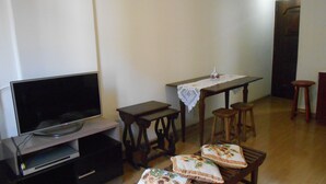 A living room with a flat-screen TV, a wooden table with a lace tablecloth, two wooden stools, and a bench with floral cushions. - Gohouse - Siqueira 703 (Rio de Janeiro)