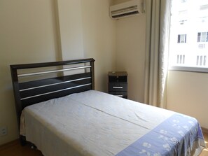 A neatly made bed with a floral bedspread, a bedside table with a remote control, and an air conditioning unit on the wall. - Gohouse - Siqueira 703 (Rio de Janeiro)